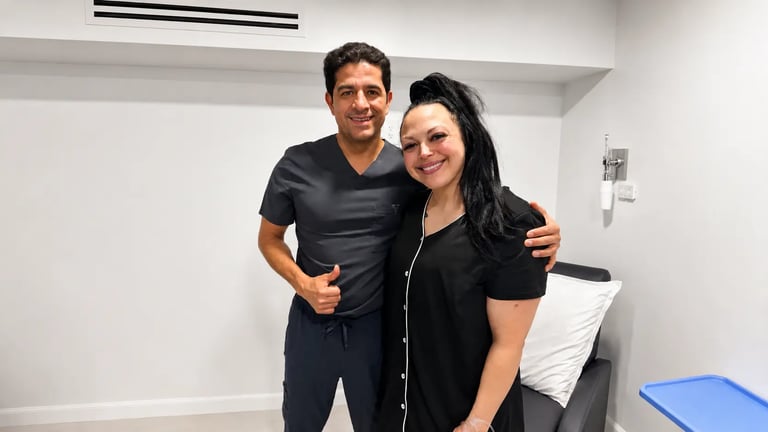 A smiling male doctor in dark grey scrubs giving a thumbs up next to a smiling female patient in a black button-up top inside a medical clinic room.