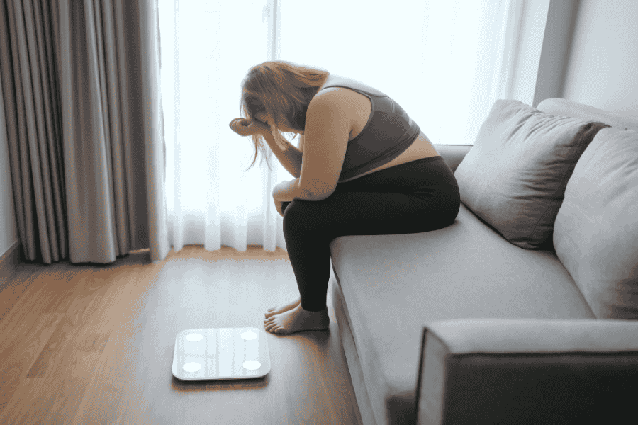 Stressed overweight woman sitting on a couch, looking at a scale on the floor, appearing frustrated. Represents the emotional impact of weight struggles and the need for bariatric solutions