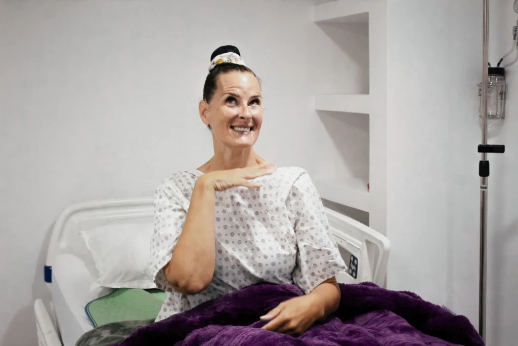 Medical Tourism in Tijuana | Affordable Surgery Packages 20 A middle-aged female patient, wearing a hospital gown, sitting up in bed with a purple blanket, smiling with a satisfied expression after her surgery at the clinic.