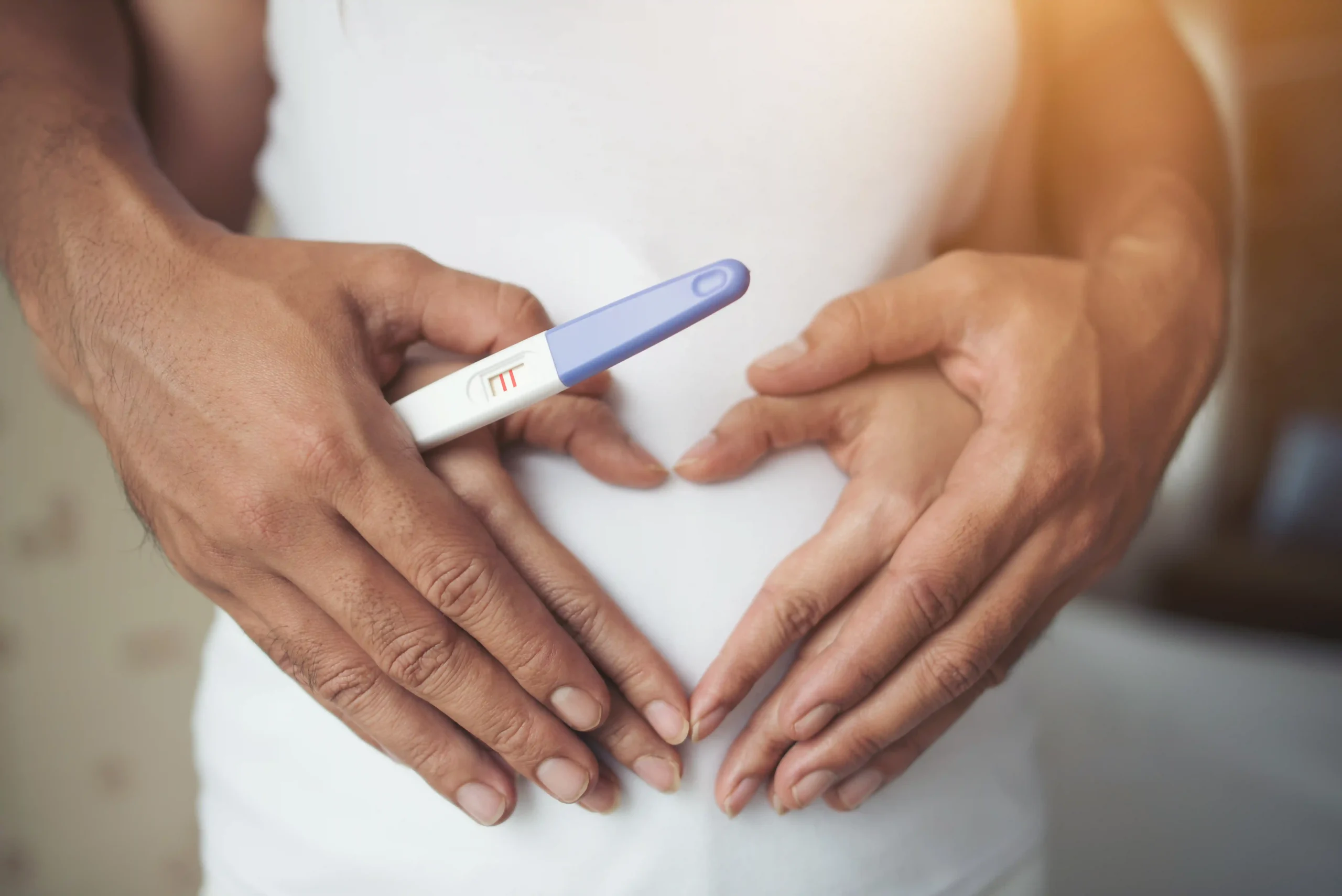 IUI Fertility in Tijuana, Mexico 11 Close-up of a couple's hands forming a heart shape over a pregnant belly while holding a positive pregnancy test.